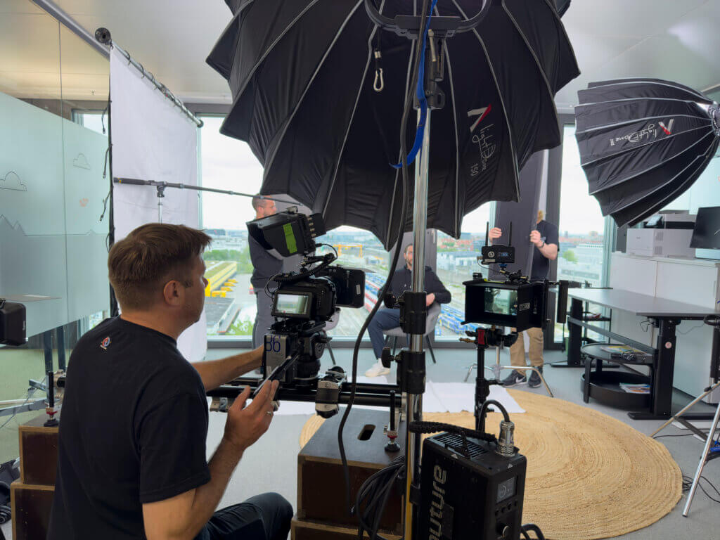 Filmteam bei der Arbeit während eines Drehs in einem modernen Büro. Kameramann bedient eine Kamera, während mehrere professionelle Lichtquellen den Raum ausleuchten.