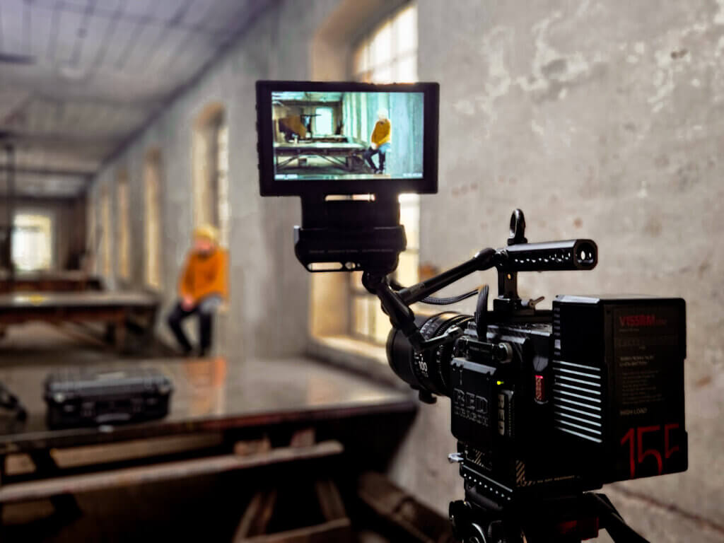 Professionelle Filmkamera mit Vorschaumonitor in einer alten Industriehalle, im Display ist ein Musiker in gelbem Pullover auf einer Werkbank sitzend zu sehen.