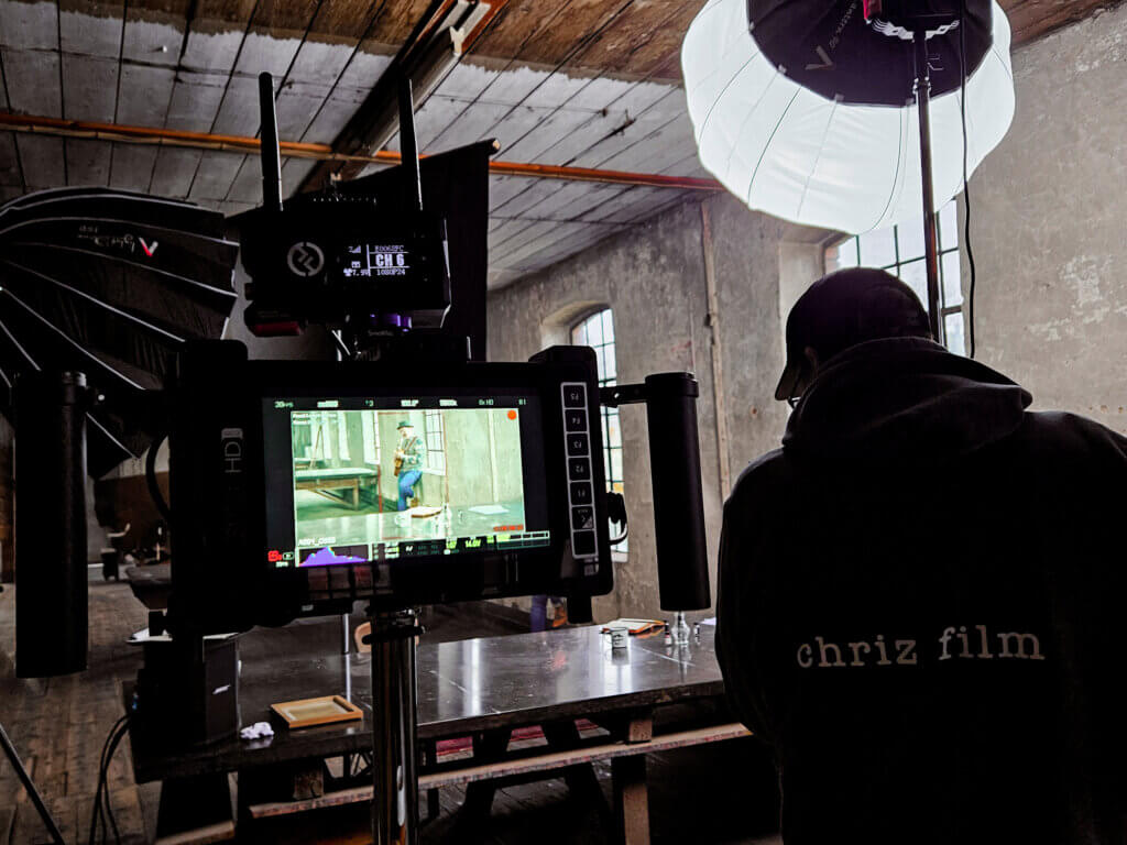 Kameramonitor zeigt eine Musiker in einer alten Industriehalle, während ein Mitglied der chriz film Crew hinter der Kamera steht und das Set ausleuchtet.