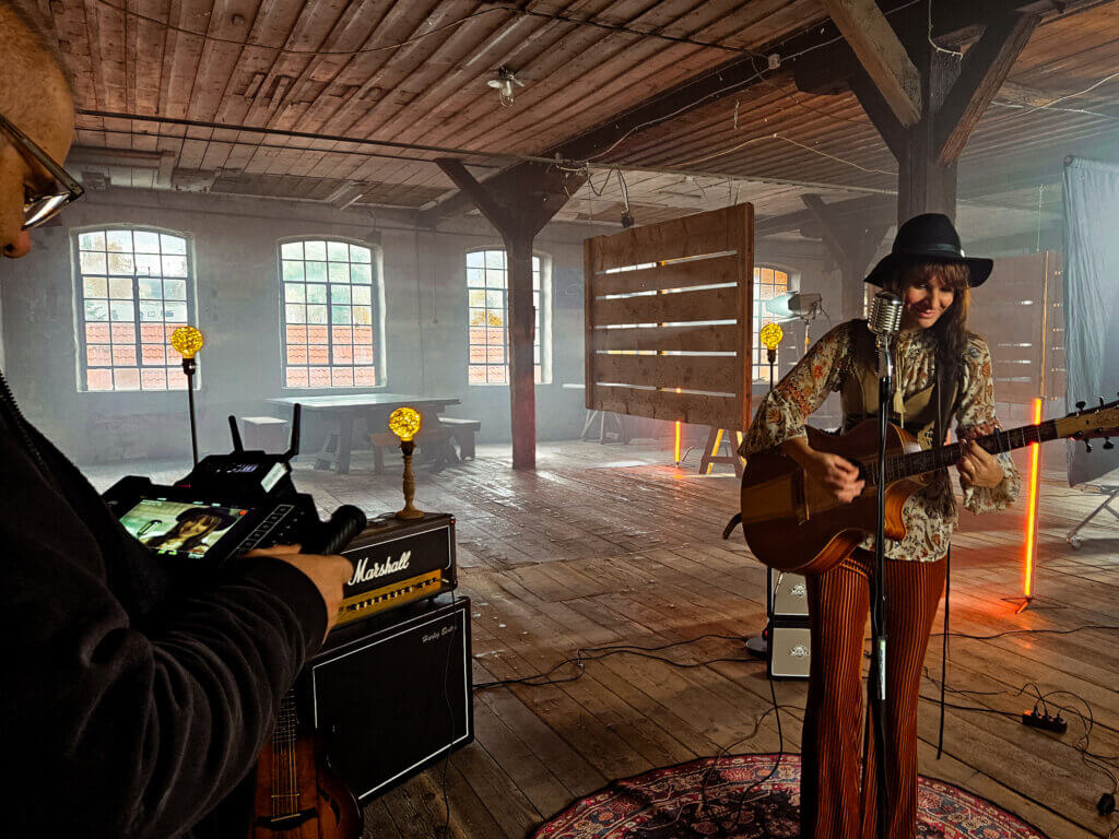Sängerin mit Gitarre tritt in einer alten Industriehalle auf, während ein Kameramann sie über einen externen Monitor filmt. Im Hintergrund warmes Bühnenlicht, Musikequipment und rustikale Holzwände.