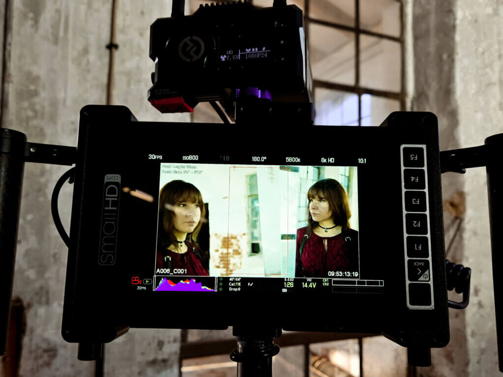 Nahaufnahme eines Kameramonitors, der zwei Porträtaufnahmen einer Musikerin zeigt. Im Hintergrund sind die Fenster und Wände der alten Industriehalle zu sehen.