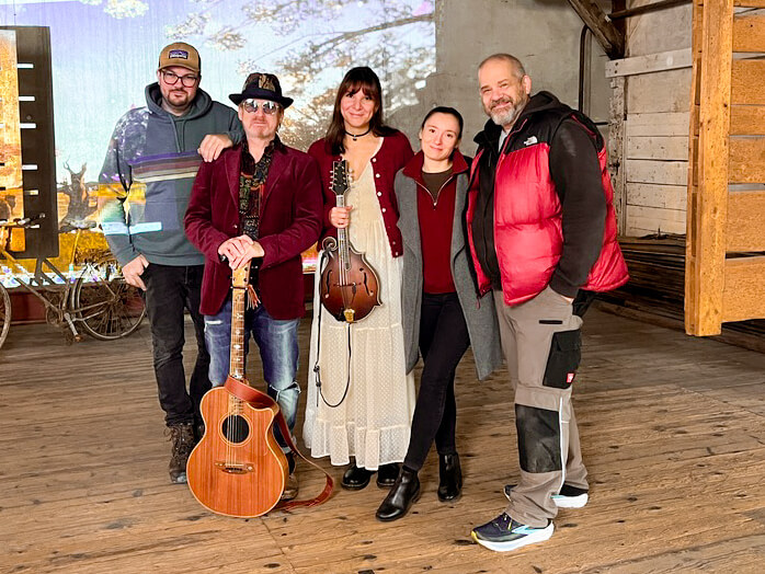 Fünf Personen stehen gemeinsam für ein Gruppenfoto in einer alten Industriehalle. Zwei Musiker halten Instrumente, eine Akustikgitarre und eine Mandoline. Die Crew lächelt in die Kamera, im Hintergrund sind Holzstrukturen und projizierte Bilder zu sehen.
